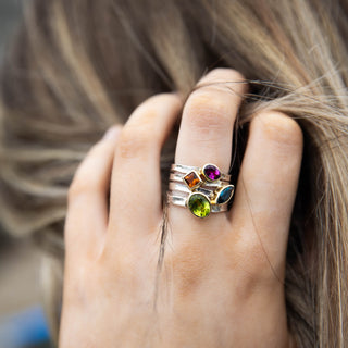 A close-up of a hand wearing a stack of three silver rings adorned with colorful gemstones, including a green peridot, a purple garnet, and a blue topaz, set against a background of brown hair.