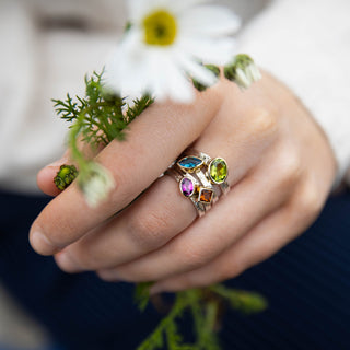 A close-up lifestyle shot of a person's hand wearing a silver ring adorned with colorful gemstones, holding a small white daisy with green foliage. The person is wearing a white long-sleeve top and a dark blue skirt.
