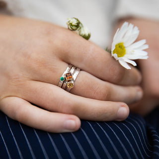 A close-up of a person's hands resting on their lap, wearing three delicate silver rings with colorful gemstone accents. One hand holds a small white daisy flower.