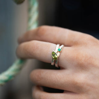 A close-up of a hand wearing a silver ring with a green marquise-cut gemstone and a small green accent stone, holding a green and white rope against a blurred background.