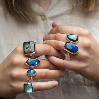 A close-up of a person wearing multiple silver rings with vibrant blue, green, and iridescent labradorite gemstones, styled with a light beige linen shirt.