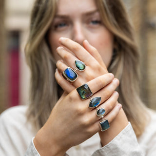 A woman wearing multiple rings with dark green and blue stones, holding her hands up to show the jewelry in a blurred outdoor setting.