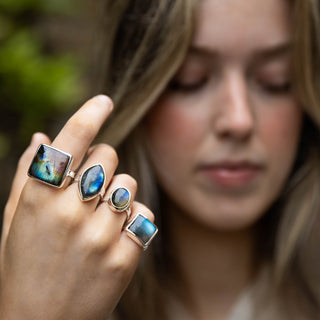 A close-up lifestyle shot of a woman wearing four silver rings with colorful  labradorite gemstones on her fingers, set against a soft-focus outdoor background.