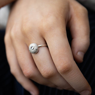 A silver ring with a textured, pale blue aquamarine circular stone is worn on a person's finger, shown in a close-up shot against dark navy pinstriped clothing.