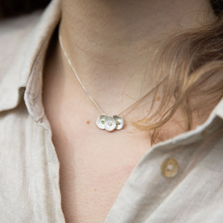 A close-up lifestyle shot of a person wearing a delicate silver necklace with three textured oval birthstone pendants, styled with a light beige linen button-up shirt.