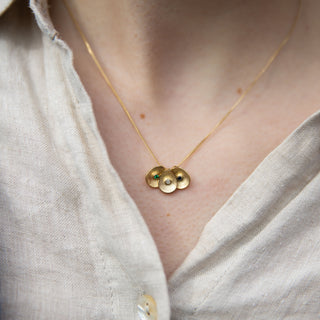 A close-up of a person wearing a delicate gold necklace with three small textured oval discs, styled over a cream linen shirt and a blue knit cardigan.