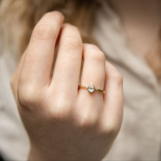 A close-up of a person's hand wearing a delicate gold ring with a pear-cut clear diamond, set against a soft-focus background of a beige top and green foliage.