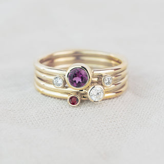 A gold multi-band stacking ring featuring a round rhodolite garnet center stone, a small round red ruby accent, and two clear round-cut diamonds, photographed on a textured off-white fabric background.
