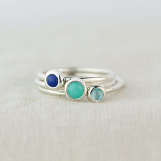 Three silver rings with green and blue gemstones sit on a light fabric background