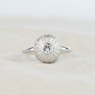 A silver ring featuring a sea urchin with a single round pale blue aquamarine gemstone in the center, resting on a soft beige fabric surface.