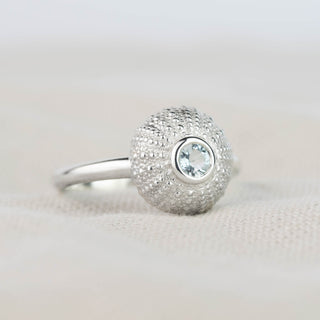 A silver ring featuring a textured, sea urchin with a central round pale aquamarine gemstone, resting on a light beige textured fabric surface.