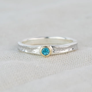 A silver band ring with a textured finish and a small round blue topaz gemstone in a gold bezel setting, resting on a light beige fabric background.