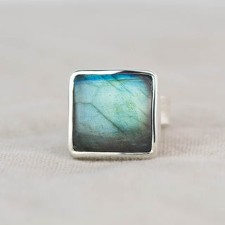 A square sterling silver ring featuring a natural labradorite stone with iridescent blue, green, and gray color shifts, set against a neutral beige textured fabric background.