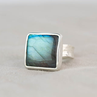 A square labradorite ring with a silver bezel and a textured silver band, resting on a neutral beige fabric background.