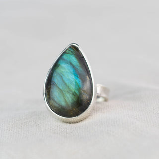 A teardrop-shaped labradorite ring with a silver bezel setting, resting on a textured off-white fabric surface. The labradorite stone displays a shimmering color shift between green, blue, and gray.