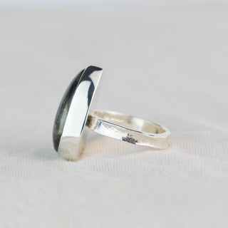 A silver ring with a large, polished, asymmetrical oval cabochon stone, shown standing on its side on a textured off-white fabric surface.