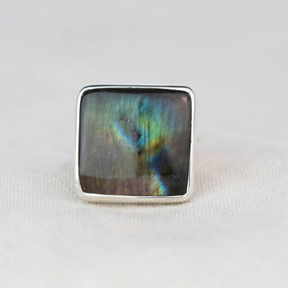 A square sterling silver ring featuring a natural iridescent labradorite stone with shimmering blue, green, and purple hues, set against a neutral light gray background.