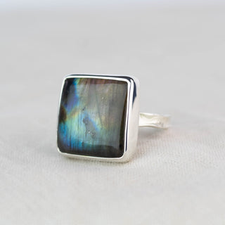 A square sterling silver ring featuring a cabochon-cut labradorite stone with a shimmering blue, green, and brown iridescent finish, set against a neutral light gray background.