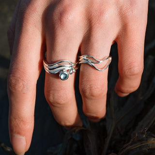 Hand wearing two silver rings with a blue gemstone, holding dark seaweed.