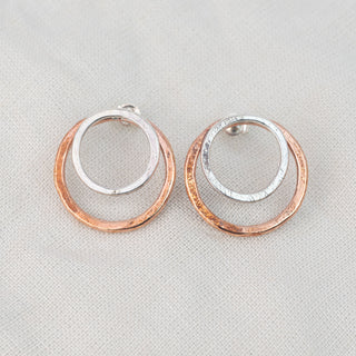 Pair of copper and sterling silver circle stud earrings resting on a wooden surface, featuring a larger copper circle with a sterling silver inner circle and lightly hammered organic textures.
