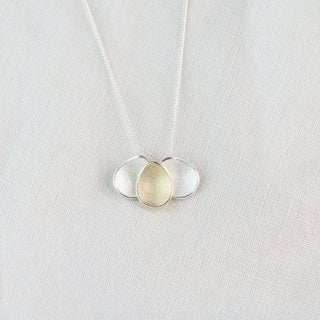 A delicate silver necklace with three interlocking teardrop-shaped pendants in polished silver and textured gold, displayed on a soft white fabric background.