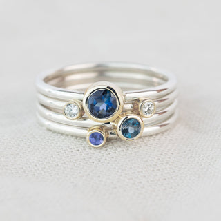 A silver ring with three stacked bands, featuring a large round blue sapphire, a smaller blue topaz, a smaller round tanzanite, and two small clear round-cut diamonds, set against a textured off-white background.