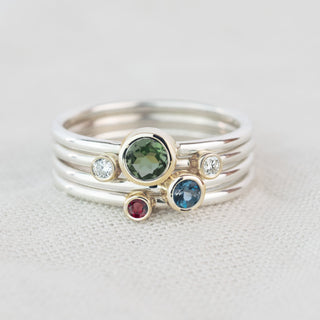 A stack of three slim silver rings on a textured off-white fabric background. The center ring features a round green tourmaline gemstone, flanked by a small clear stone and a blue topaz gemstone, with a separate gold-accented ring below featuring a small red ruby.