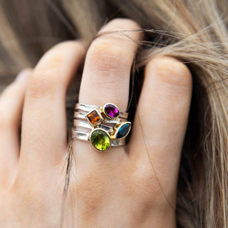  Close-up of four Storybook stacking rings worn on a finger, featuring coloured gemstones in gold settings including London Blue Topaz, Peridot, Rhodolite Garnet and Citrine, by Alison Moore Designs