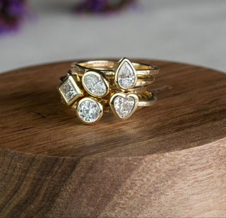 A stack of gold rings with diamond gemstones in pear, round, and square settings, displayed on a round wooden pedestal with a soft-focus background.
