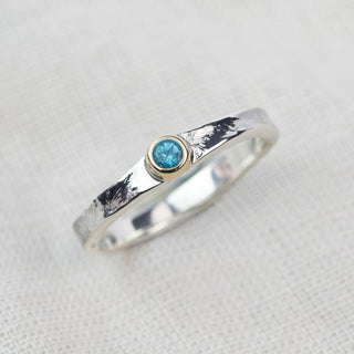 A silver band ring with a textured, hammered finish and a small bezel-set round blue topaz gemstone, displayed on a light beige linen fabric background.
